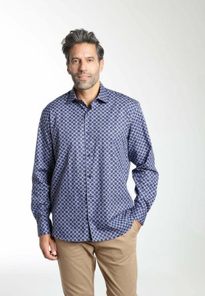 Homme debout portant une chemise à boutons bleue à motifs et un pantalon beige, une main dans la poche, sur fond blanc.