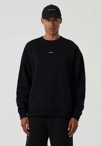 Felpa nera oversize con scollo rotondo e un piccolo logo bianco. Indossata con pantaloni neri abbinati e un cappellino da baseball nero.
