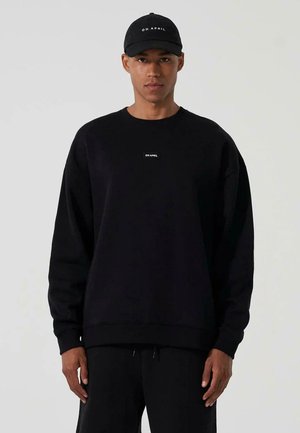 Svart oversized hettegenser med rund hals og et lite hvitt logo-patch. Kombinert med matchende svarte bukser og en svart baseballcaps.