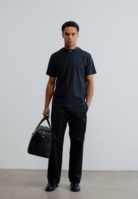 Μαύρο κοντομάνικο μπλουζάκι με μίνιμαλ σχεδιασμό, συνδυασμένο με άνετο μαύρο παντελόνι. Το άτομο κρατά μια μεγάλη μαύρη τσάντα duffel με μεταλλικές λεπτομέρειες.