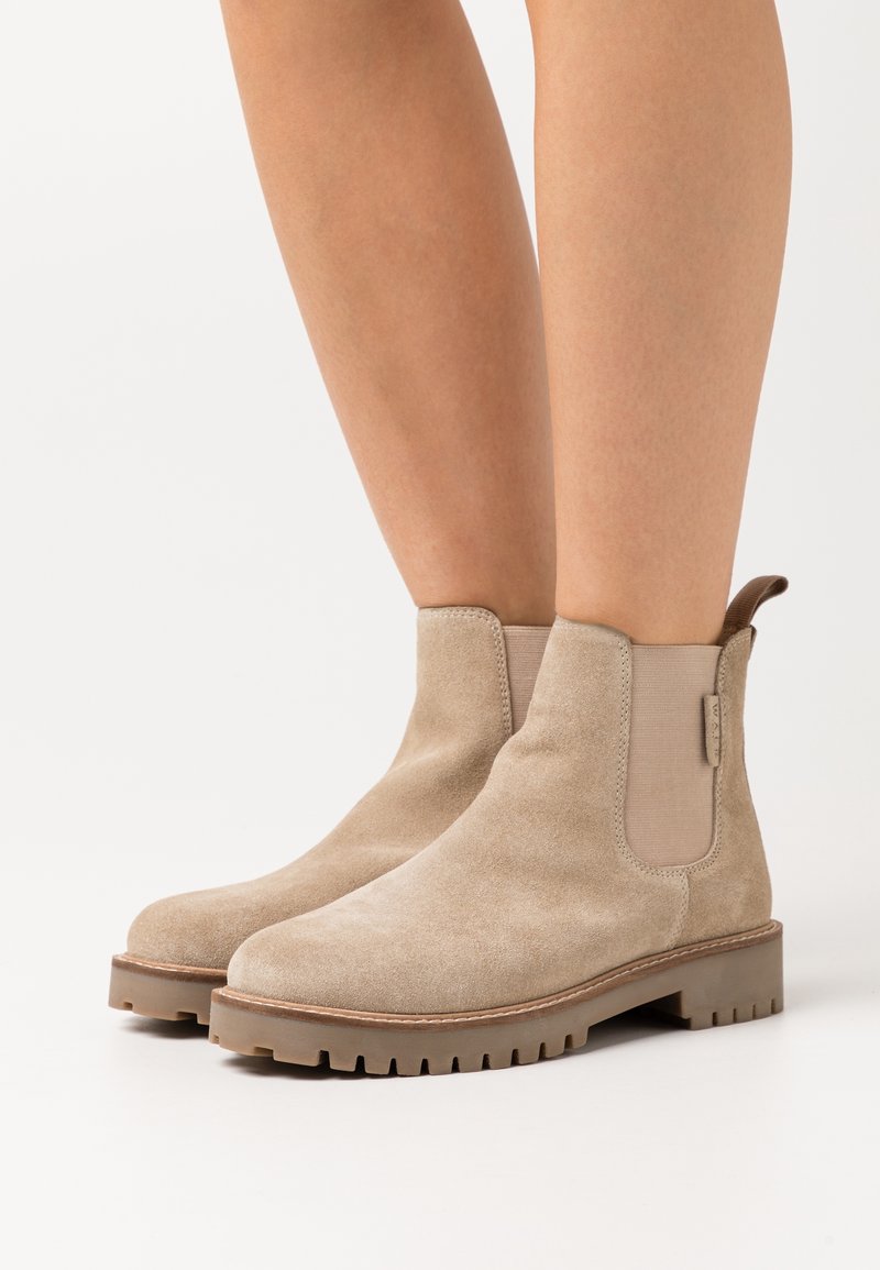 Bottines beige en daim avec panneaux latéraux élastiques, bouts arrondis et semelles en caoutchouc motifées pour une meilleure traction. Comprend une languette à l'arrière.