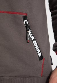 Felpa grigia con cuciture rosse, dotata di tasca con zip e un'etichetta logo in bianco e nero. Texture morbida e dettaglio a coste sull'orlo.