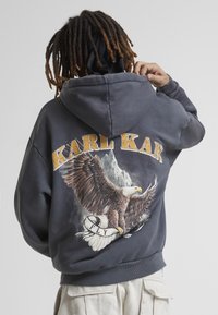 Felpa con cappuccio grigia che presenta una grande grafica di un'aquila e "KARL KANI" in giallo sopra. Tessuto di notevole consistenza con una vestibilità rilassata.