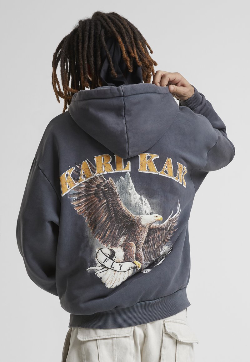 Felpa con cappuccio grigia che presenta una grande grafica di un'aquila e "KARL KANI" in giallo sopra. Tessuto di notevole consistenza con una vestibilità rilassata.