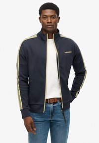 Veste zippée marine en tissu doux, ornée de rayures latérales vert clair, d'un col montant et d'un petit logo doré sur la poitrine.