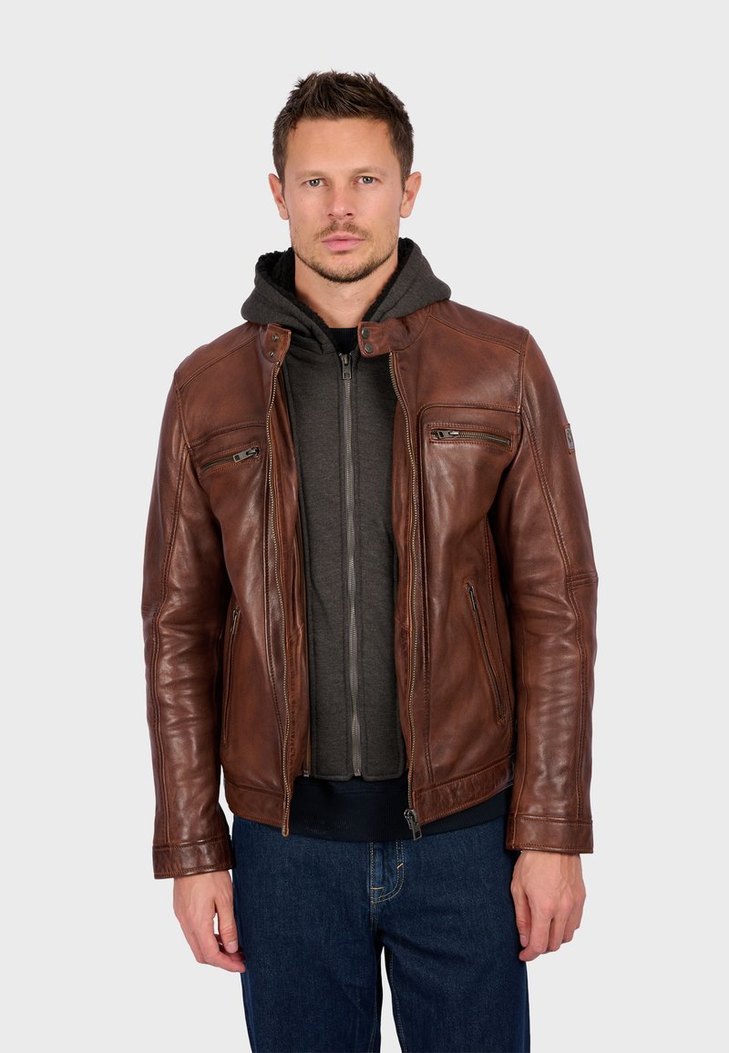 Man in een bruin lerenjas over een donkergrijze hoodie met rits en blauwe spijkerbroek, staand tegen een effen lichtgrijze achtergrond.