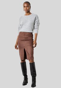 Femme portant un pull en tricot gris, une jupe marron à mi-mollet avec une fente à l'avant, des bottes noires jusqu'aux genoux et des boucles d'oreilles créoles sur un fond gris.