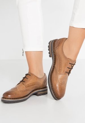 Braune Brogue-Lederschuhe mit schwarzen Schnürsenkeln, getragen zu weißen Hosen, mit detaillierter Nahtführung und Gummisohlen.