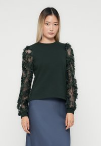 Donna con capelli lisci che indossa un top verde scuro con maniche trasparenti a fantasia floreale e una gonna blu, in piedi davanti a uno sfondo semplice.
