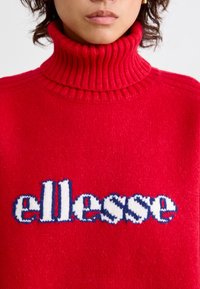 Rode gebreide coltrui met ribbeltextuur, met centraal op de voorkant het woord "ellesse" in wit en blauw.