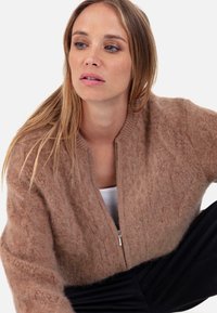 Brauner, flauschiger Strickcardigan mit Reißverschluss vorne und geripptem Kragen. Mit Zopfmuster und lockerer Passform. Wird über einem weißen Oberteil getragen.