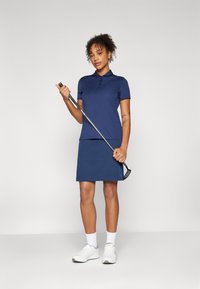 Camisa polo azul-marinho com colarinho e botões, combinada com uma saia de golfe azul coordenada. Ténis brancos e um taco de golfe na mão.