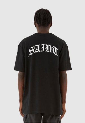 Svart bomull T-skjorte med stor hvit tekst "SAINT" på baksiden. Oversized passform med korte ermer og rund hals. Glatt tekstur.