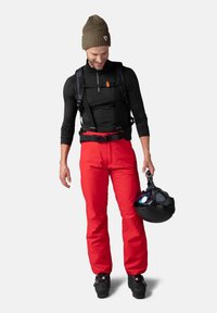 Rote Skihose, schwarzes Langarmoberteil, brauner Strickmütze und schwarze Stiefel. Die Person hält einen schwarzen Helm mit blauem Licht.