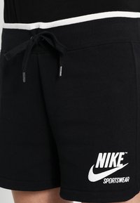 Pantalones cortos Nike negros con una cinturilla elástica blanca, cordón ajustable y estampado del logo blanco en la parte inferior izquierda. Textura de tela suave.