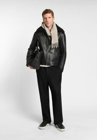 Schwarze Lederjacke mit einem Pelzkragen, beiger Schal, schwarze Hose und schwarze Sneaker mit weißen Sohlen. Hält eine strukturierte schwarze Tasche.