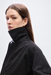 Νέα γυναίκα με μακριά καστανά μαλλιά που φοράει μαύρο μπουφάν με ανασηκωμένο γιακά, κοιτάζοντας πλάγια μπροστά σε απλό ανοιχτό φόντο.