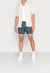 Vaaleanbeige lyhythihainen paita, valkoinen tanktop, raidalliset laivastonsiniset shortsit käännetyllä helmalla, valkoiset tennarit, seisoo neutraalilla taustalla.