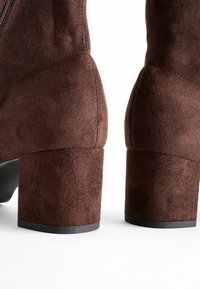 Bottes de cheville en daim marron avec un talon bloc, texture lisse et un détail de fermeture éclair sur un côté. Les talons sont épais et de forme carrée.