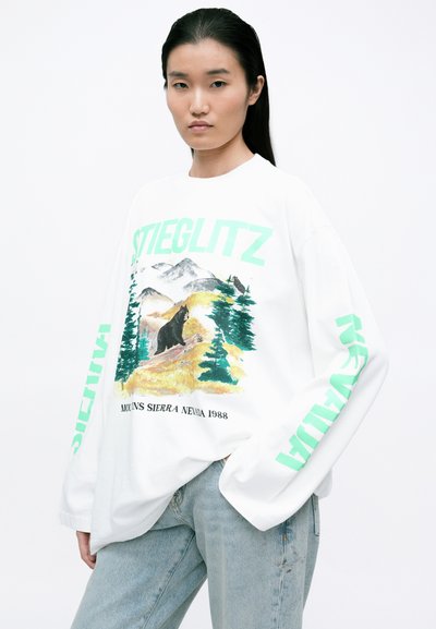 Biely sweatshirt s dlhým rukávom s farebnou grafikou medveďa a hory. Text "STIEGLITZ" v zelenej farbe pozdĺž rukávov. Mäkko, pohodlný strih.