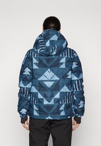 Giacca a vento blu trapuntata con un motivo geometrico in tonalità più scure. Include un cappuccio e polsini elastici per una vestibilità aderente.