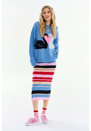 Kobieta o długich blond włosach, ubrana w niebieski sweter z wzorem łabędzia i serca, kolorową spódnicę midi w paski, różowe skarpetki i różowe trampki.