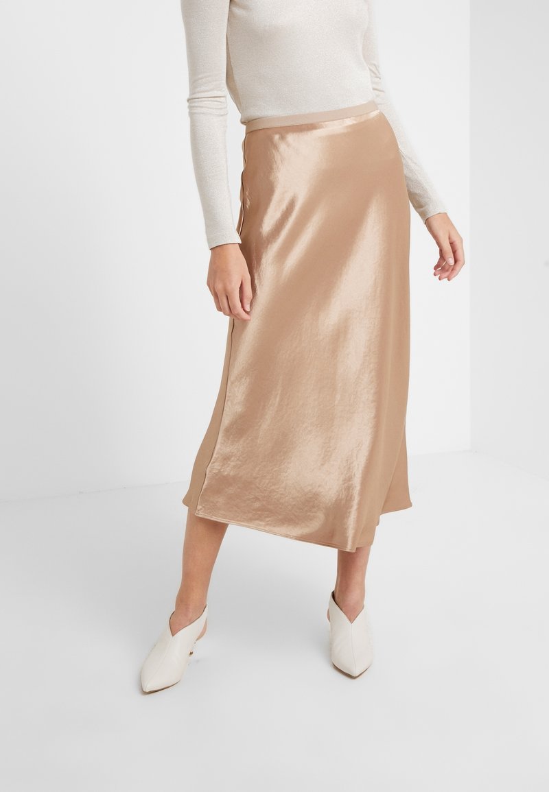 Nude satynowa spódnica midi o gładkiej teksturze, z delikatnym połyskiem i prostym, zwiewnym krojem. Połączona z beżowymi szpilkami w kształcie migdałów.