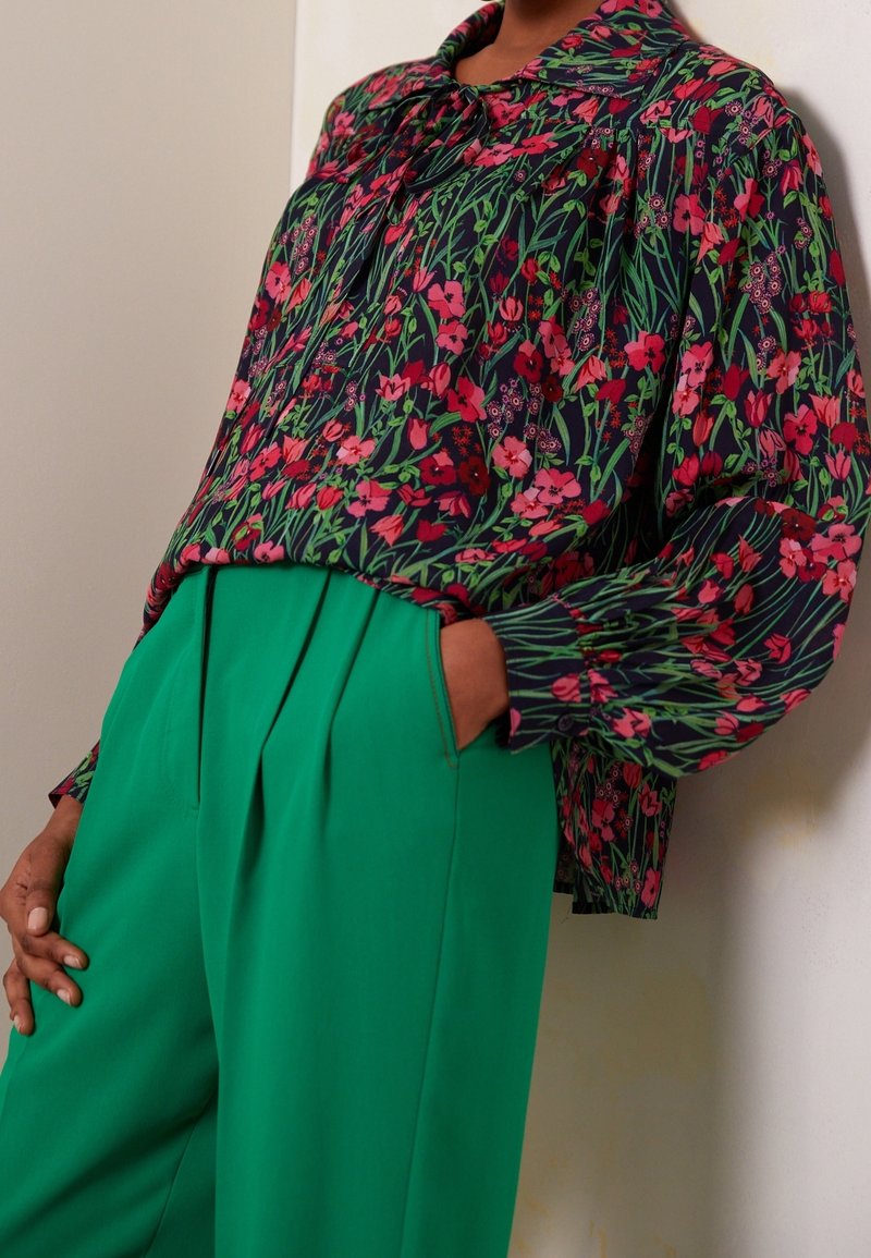 Personne portant un chemisier à manches longues à fleurs roses et rouges et un pantalon taille haute vert vif avec des plis et des poches.