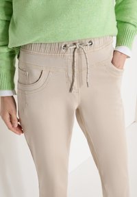 Pantalon beige clair à taille à cordon porté avec un pull vert, une main dans la poche et l'autre détendue le long du corps.