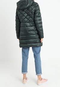 Tummanvihreä, tilkkuvuorattu puffer-takki hupulla. Siinä on vartalonmyötäinen vyötärö, pitkät hihat, ja se on yhdistetty vaaleisiin farkkuihin ja vaaleanpunaisiin lenkkareihin.