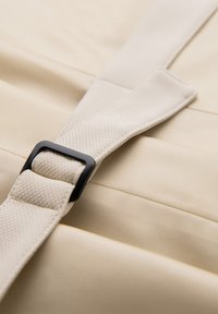 Gros plan d'une sangle en tissu beige avec une boucle rectangulaire noire sur un fond de matière couleur crème.