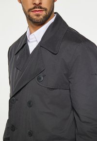 Trench coat nero doppiopetto con grandi bottoni, revers larghi e una texture liscia. Presenta una vestibilità su misura e dettagli di cucitura visibili.