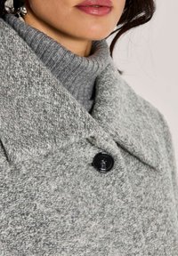 Vrouw in een gestructureerde grijze woljas over een ribbelachtige grijze coltrui, met een zichtbare grote zwarte knoop en roze lippen.