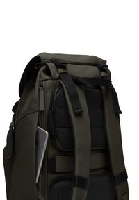 Sac à dos vert foncé avec des bretelles réglables, panneau en maille noire texturée, poche externe et un smartphone gris visible rangé à l'intérieur.