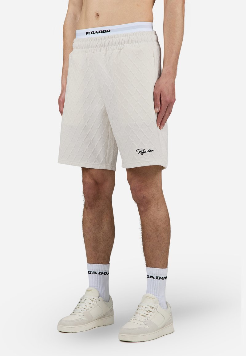 Shorts off-white avec texture en losanges matelassés, taille élastique et logo de la marque brodé. Associés à des baskets blanches et des chaussettes.