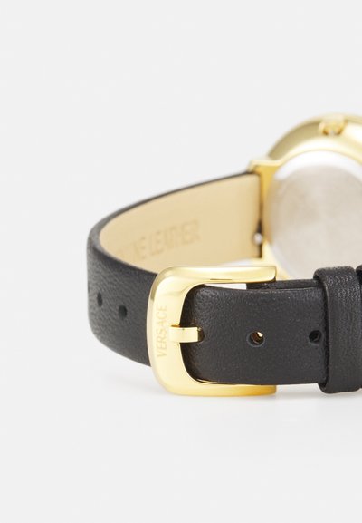 Curea de ceas din piele neagră cu un finisaj texturat, cu o cataramă din metal de culoare aurie, gravată cu „VERSACE”. Fața ceasului circular este vizibilă.