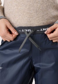 Navy waterproof pants with a gray elastic waistband and drawstring. Smooth, glossy texture with branding on the waistband.