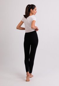 Zwarte, strakke leggings met een elastische tailleband, gecombineerd met een eenvoudig wit T-shirt. Model loopt op blote voeten, richt zich weg van de camera en toont een eenvoudig ontwerp.