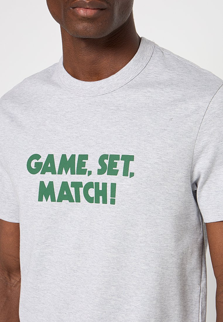 Graues Baumwoll-T-Shirt mit fettem grünen Text, der auf der Vorderseite "GAME, SET, MATCH!" liest. Runder Halsausschnitt und kurze Ärmel.