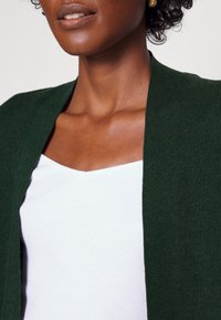 Ciemnozielony sweter z dzianiny z otwartym przodem, w połączeniu z białym podkoszulkiem o prążkowanej fakturze. Skupienie na fakturze i warstwowych elementach projektu.