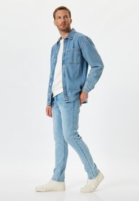 Chaqueta de mezclilla de un azul claro sobre un suéter blanco, combinada con jeans de azul claro y zapatillas crema. Presenta un bolsillo en el pecho y cierre de botones.
