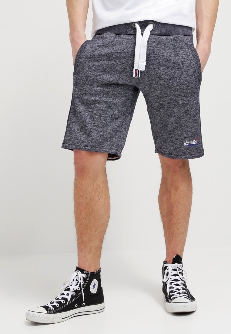 Homme portant un short gris foncé à cordon de serrage et des baskets montantes noires avec des lacets blancs, debout les mains dans les poches.