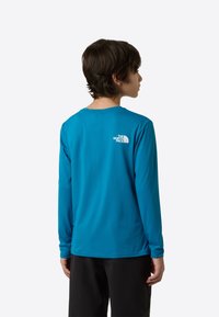 Mladinska majica z dolgimi rokavi iz svetlo modrega blaga, z okroglim izrezom in majhnim belim logotipom na hrbtu. Gladek tekstura, sproščen kroj.