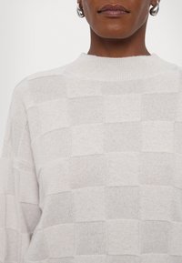 Maglione grigio chiaro con un motivo a scacchi testurizzato, colletto a coste e tessuto morbido con una leggera lucentezza.