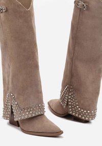 Bottines en daim taupe avec des embouts pointus, accentuées par des clous argentés et un détail découpé unique sur la tige. Semelles blocs pour plus de stabilité.