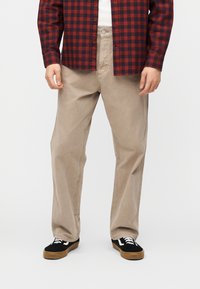 Άνδρας που φοράει μπεζ παντελόνι άνετης γραμμής, μαύρα και καφέ αθλητικά παπούτσια και κόκκινο-μαύρο καρό πουκάμισο, στέκεται μπροστά σε λευκό φόντο.