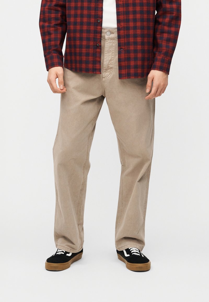 Άνδρας που φοράει μπεζ παντελόνι άνετης γραμμής, μαύρα και καφέ αθλητικά παπούτσια και κόκκινο-μαύρο καρό πουκάμισο, στέκεται μπροστά σε λευκό φόντο.