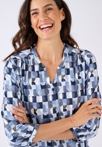 Camicia button-up di colore azzurro chiaro con motivi geometrici in blu navy, bianco e nero. Presenta un collo tondo e maniche a tre quarti.