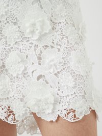 Tissu en dentelle blanche avec des appliqués floraux en relief et un ourlet festonné. Des motifs floraux complexes créent une surface texturée.