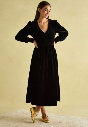 Schwarzes Samtkleid mit langen Ärmeln, V-Ausschnitt, gerafftem Oberteil und fließendem Rock, kombiniert mit goldenen Mary Jane Schuhen. Glatte Textur.